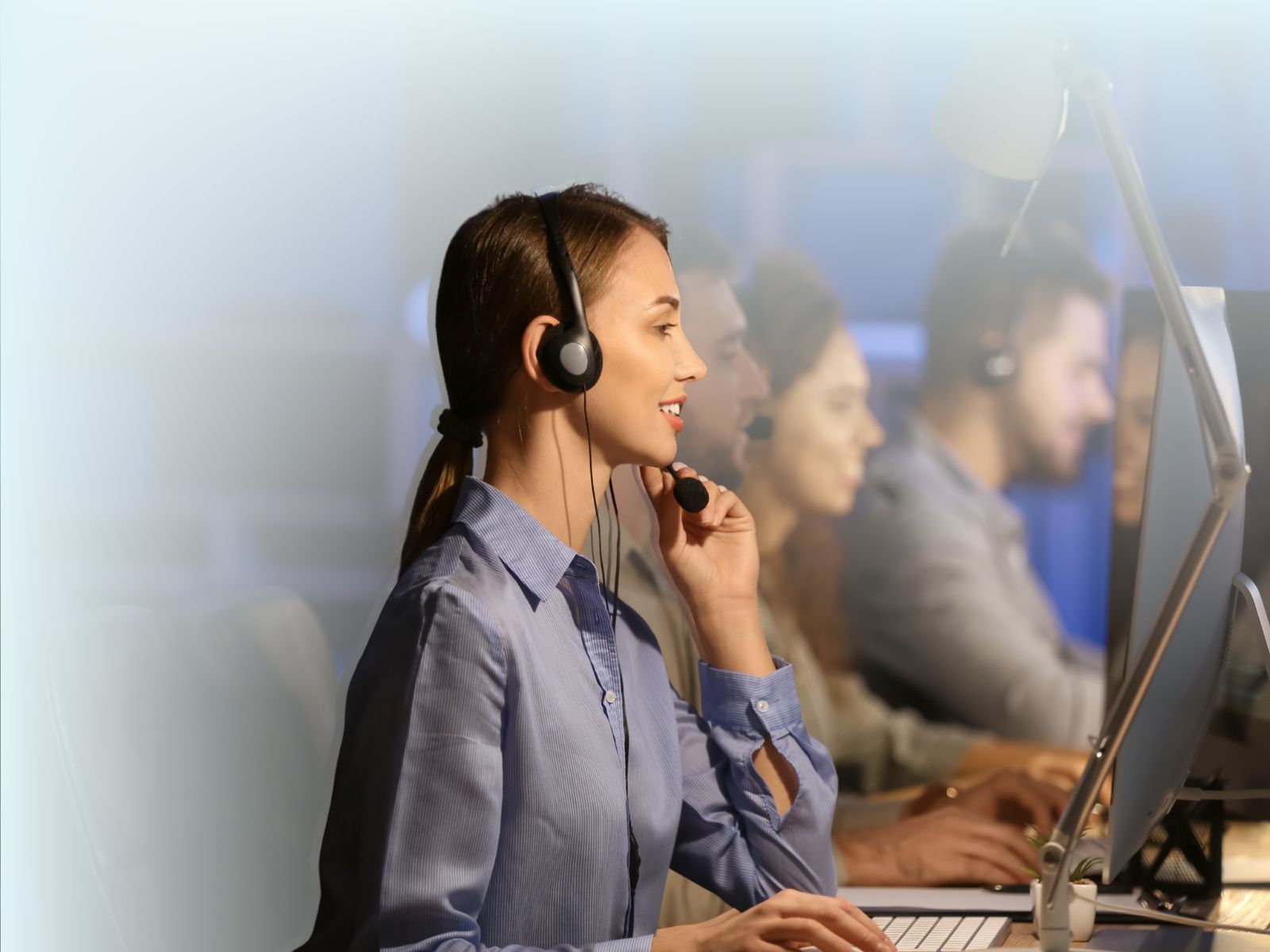 Agente de call center bilingüe sonriendo y usando un headset mientras trabaja en su estación, rodeado de otros agentes en un centro de contacto.