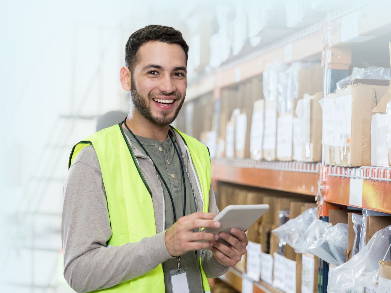Joven almacenista o auxiliar logístico sonriendo con chaleco de seguridad reflectante, revisando datos en una tablet en el pasillo de un almacén.