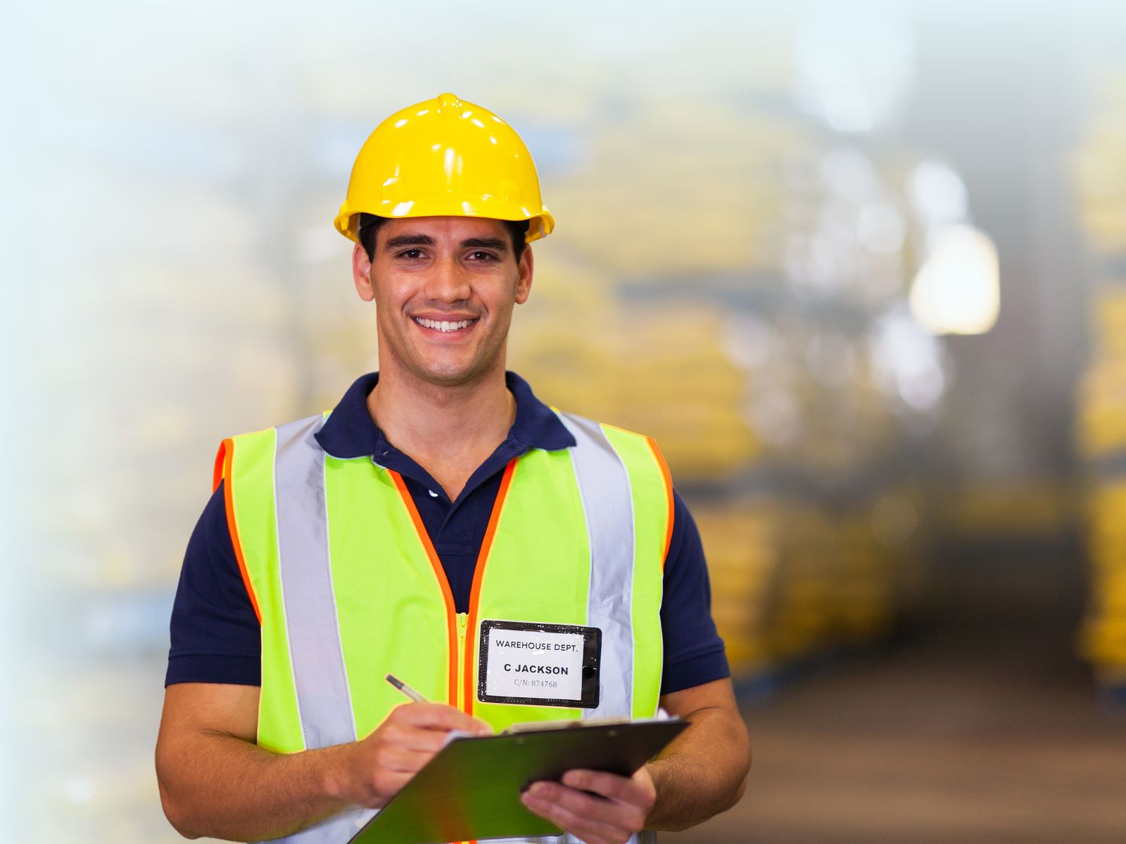 Supervisor o trabajador de almacén sonriente con casco de seguridad amarillo, chaleco reflectante y portapapeles, en un entorno industrial.